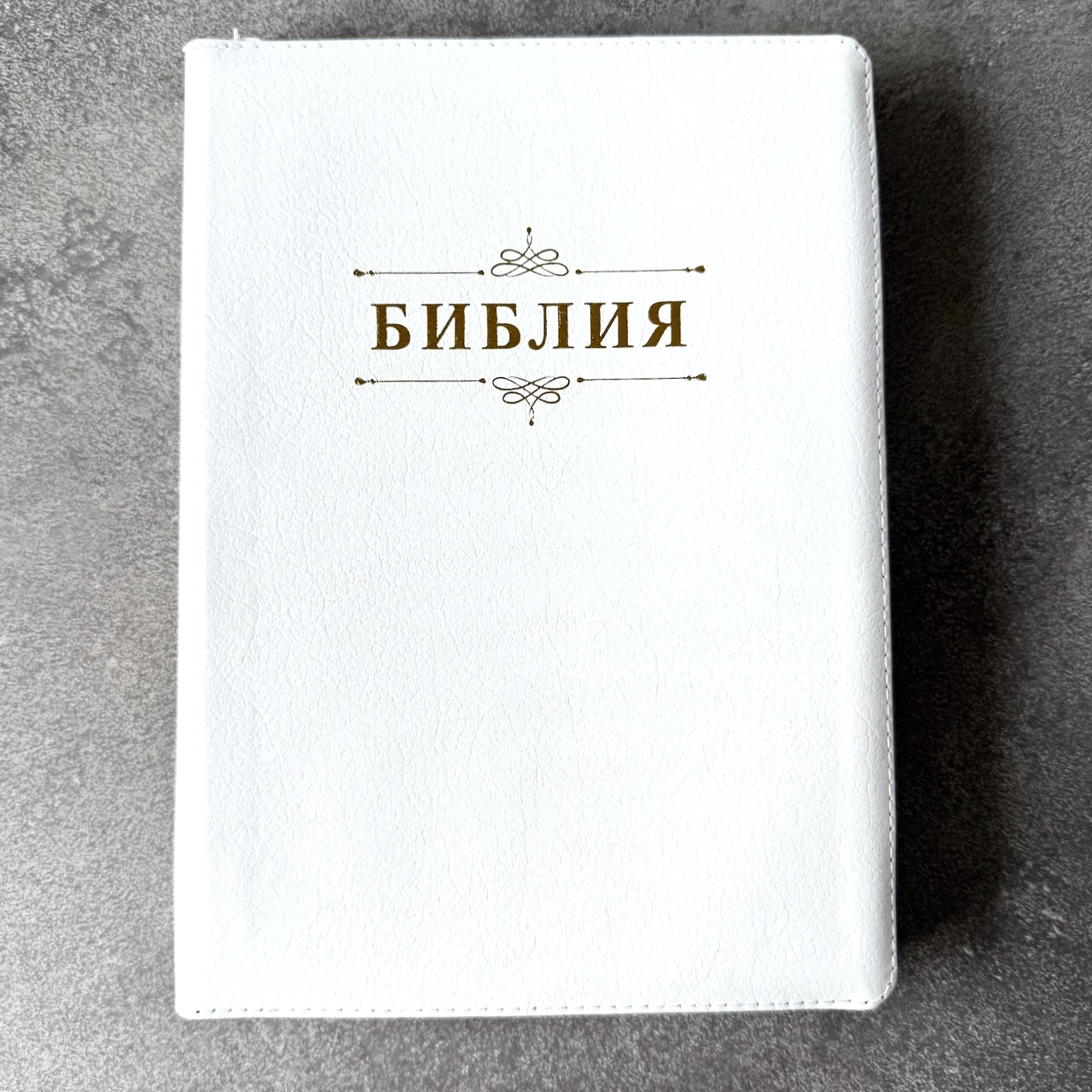 Дисконт. Маленькая серая точка на лицевой обложке, слева от центра. Библия 076zti код 25076-16, дизайн "слово Библия", кожаный переплет на молнии с индексами, цвет белый пятнистый, размер 170x243 мм