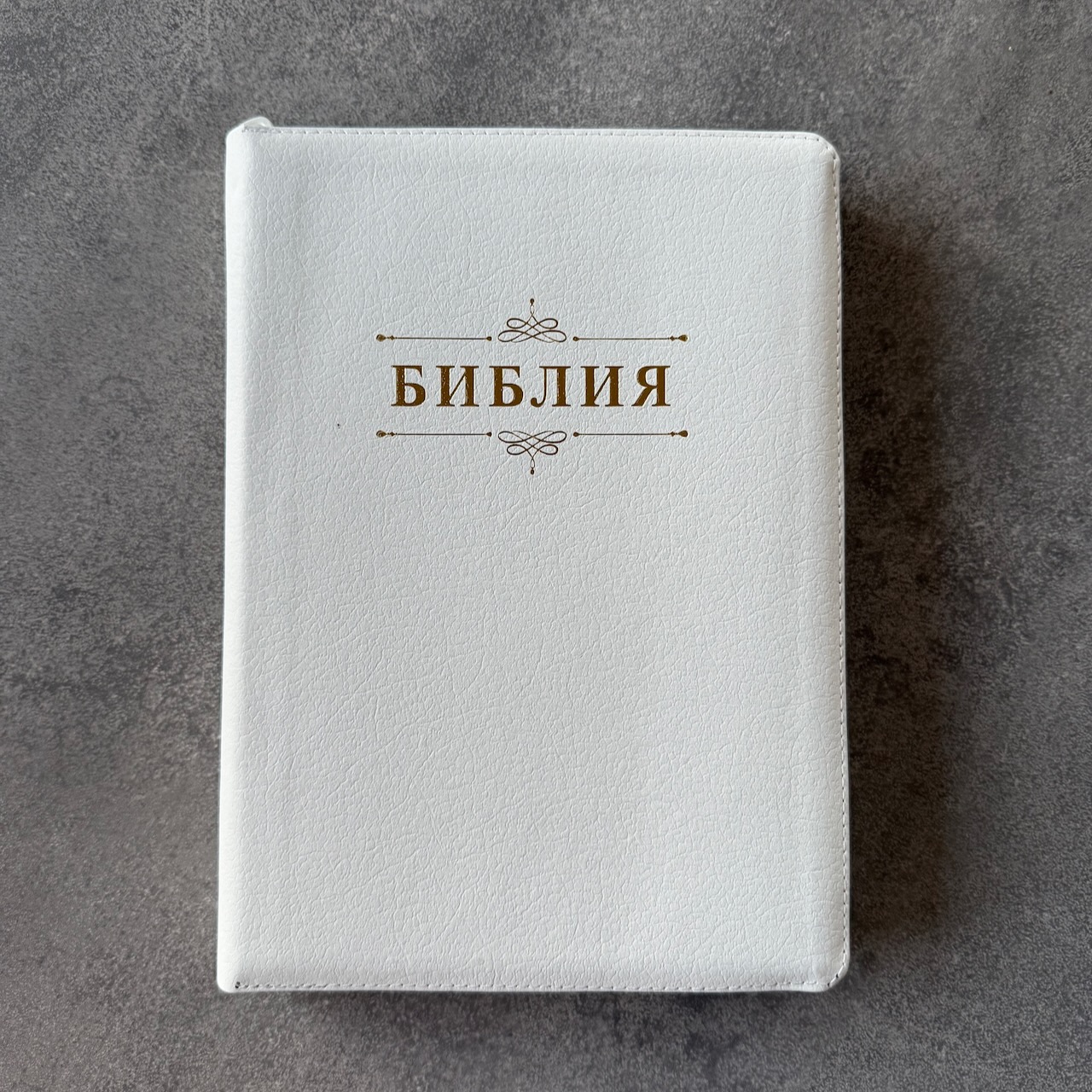 Дисконт. Маленькая серая точка на лицевой обложке, слева от центра. Библия 076zti код 25076-16, дизайн "слово Библия", кожаный переплет на молнии с индексами, цвет белый пятнистый, размер 170x243 мм